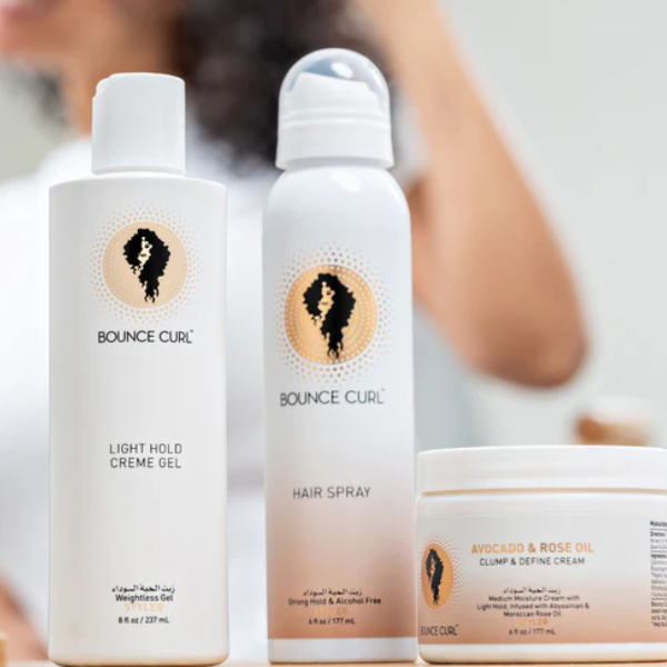 Drie Bounce Curl haarproducten op een aanrecht: Light Hold Creme Gel in een witte fles (8 fl oz/237 ml), Hair Spray in een witte spuitbus (8 fl oz/177 ml), en Avocado & Rose Oil Clump & Define Cream in een pot (8 fl oz/237 ml). Op de achtergrond is vaag een persoon met krullend haar te zien, wat benadrukt dat de producten bedoeld zijn voor de verzorging van krullend haar